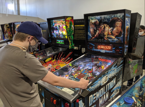 A person plays a pinball machine titled "Demolition Man" in an arcade filled with various games.