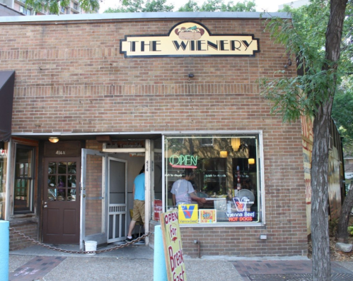 A brick building with a sign reading "The Wienery" and an open door, featuring a colorful "Open" sign in the window.