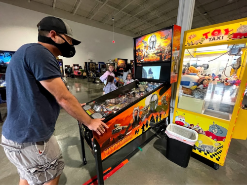 A person plays a Mandalorian pinball machine in an arcade, with a claw machine and other players in the background.