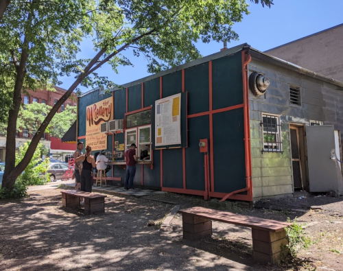 A small, colorful food stand with outdoor seating, surrounded by trees and a parking area on a sunny day.