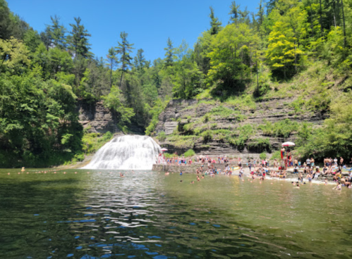 A sunny day at a waterfall with people swimming and relaxing by the water, surrounded by lush green trees and rocky cliffs.