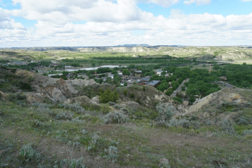 A panoramic view of a lush valley with a small town, surrounded by rolling hills and a cloudy blue sky.