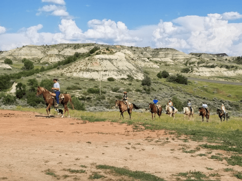 A group of horseback riders trails through a scenic landscape of hills and blue skies.