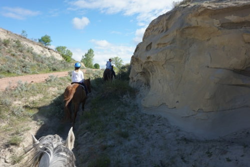 Two riders on horseback navigate a sandy trail beside a rocky cliff under a blue sky.