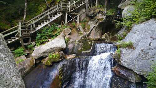 A serene waterfall cascades over rocks, with a wooden staircase leading through lush greenery in the background.