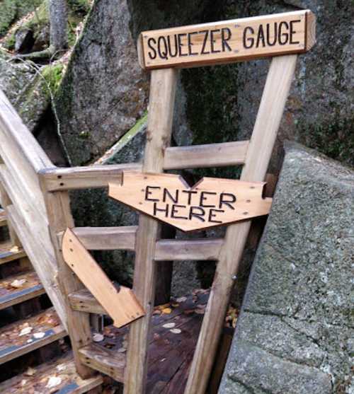 Wooden sign with arrows reading "Squeezer Gauge" and "Enter Here," positioned near a set of stairs in a natural setting.