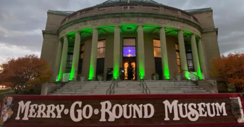 Exterior of the Merry-Go-Round Museum, featuring green lights and a sign in front, against a cloudy evening sky.