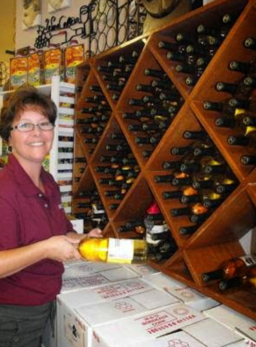 A woman in a maroon shirt smiles while holding a bottle of wine in a store with wine racks in the background.