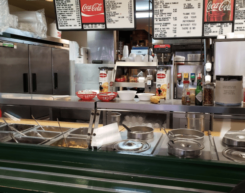 A restaurant counter with food trays, condiments, and a menu board in the background.