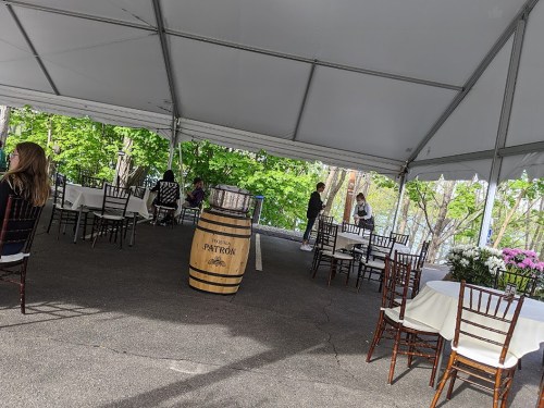 A tented outdoor space with empty tables, chairs, and a decorative barrel, surrounded by greenery.