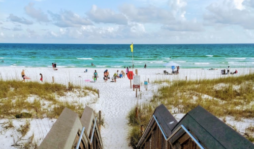 A sandy beach with turquoise water, people relaxing, and beach chairs under a blue sky with scattered clouds.