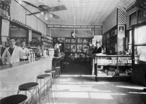 Historic interior of a vintage café, featuring a bar, shelves of goods, and two staff members in white aprons.