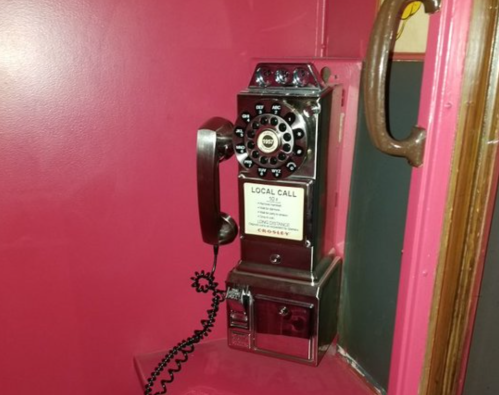 A vintage payphone mounted on a bright pink wall, featuring a rotary dial and a coin slot.