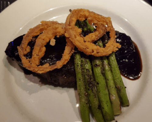 A plated dish featuring a steak topped with onion rings, served with asparagus and a rich sauce.