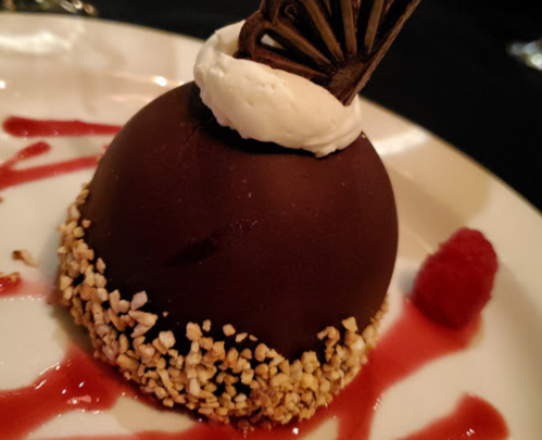 A chocolate dome dessert topped with whipped cream, surrounded by raspberry sauce and a raspberry garnish.