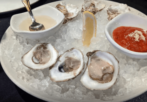 A plate of fresh oysters on ice, served with lemon wedges and two dipping sauces.