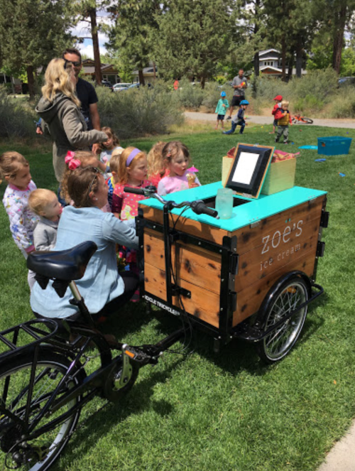 A group of children eagerly gathers around a colorful ice cream cart in a park on a sunny day.