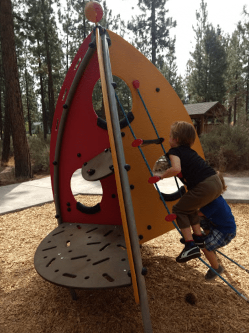 Two children play on a colorful climbing structure in a park surrounded by trees.