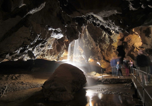 A dimly lit cave with a waterfall cascading over rocks, and visitors observing the natural beauty.