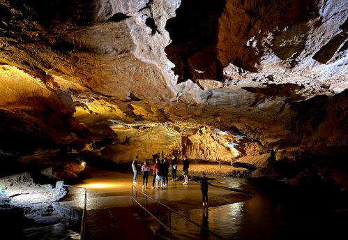 A group of people exploring a large, illuminated cave with rocky formations and a water surface.