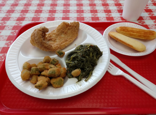 A plate with fried fish, cooked greens, fried okra, and two breadsticks on a red tray with a checkered tablecloth.
