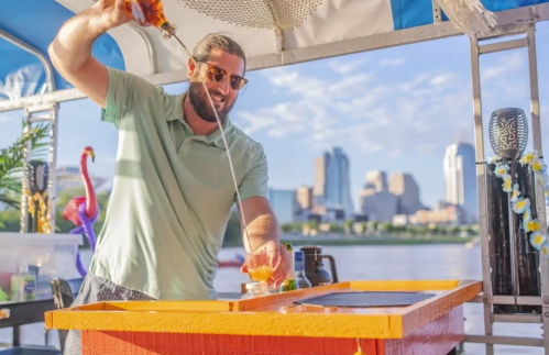 A man pours a drink from a bottle at a colorful bar on a boat, with a city skyline in the background.