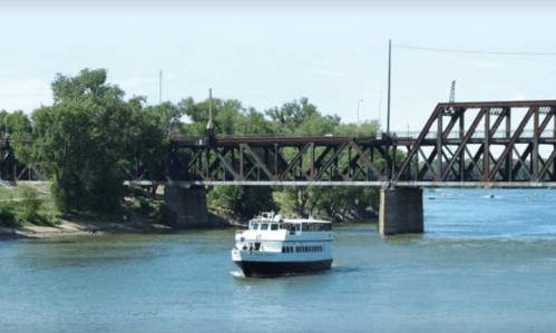 A boat navigates a river near a metal railway bridge surrounded by greenery.