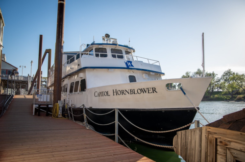 A boat named "Capitol Hornblower" docked at a wooden pier on a calm river.