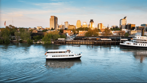 A boat glides on a river with a city skyline in the background, featuring buildings and greenery along the shore.