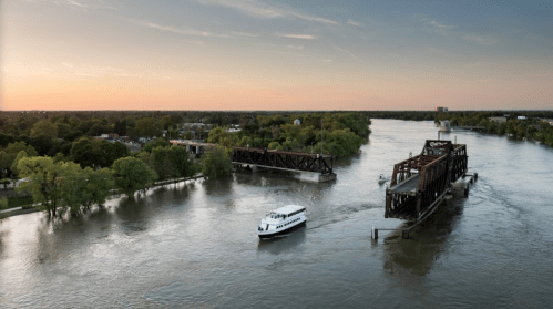 A boat navigates a river near a bridge at sunset, surrounded by lush greenery and calm waters.