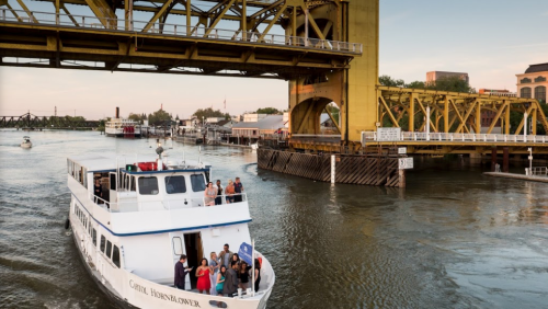 A riverboat with passengers sails under a yellow bridge, surrounded by water and buildings along the shore.