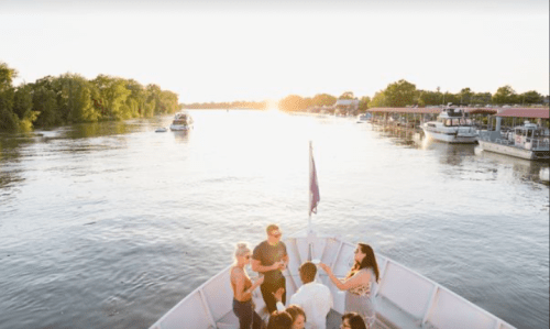 A group of people enjoying a sunset on a boat, with a calm river and docks in the background.