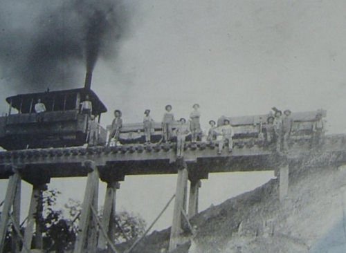 Black and white photo of a steam train on a wooden bridge with workers posing nearby.