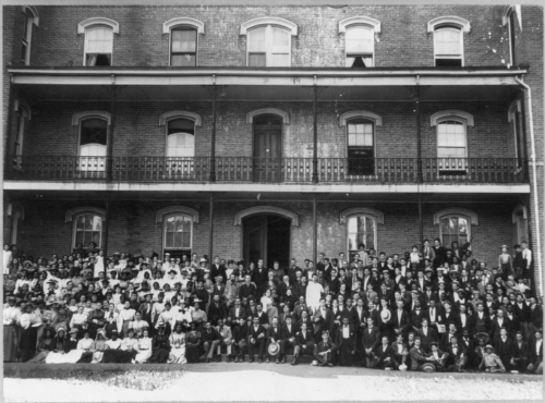 A large group of people in formal attire gathered in front of a historic brick building with multiple balconies.
