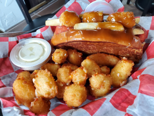 A hot dog topped with fries and sauce, served with a side of crispy tater tots and dipping sauce in a red-checkered basket.