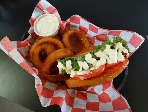 A basket with a hot dog topped with lettuce and tomatoes, served with onion rings and a small cup of sauce.