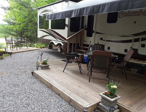 A cozy outdoor space with a gravel path, wooden deck, chairs, and a camper surrounded by greenery.
