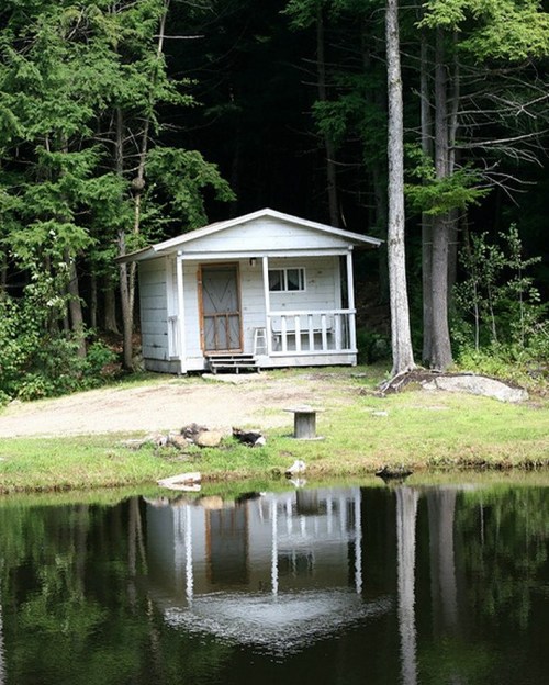 A small white cabin by a calm pond, surrounded by lush green trees, reflecting in the water.