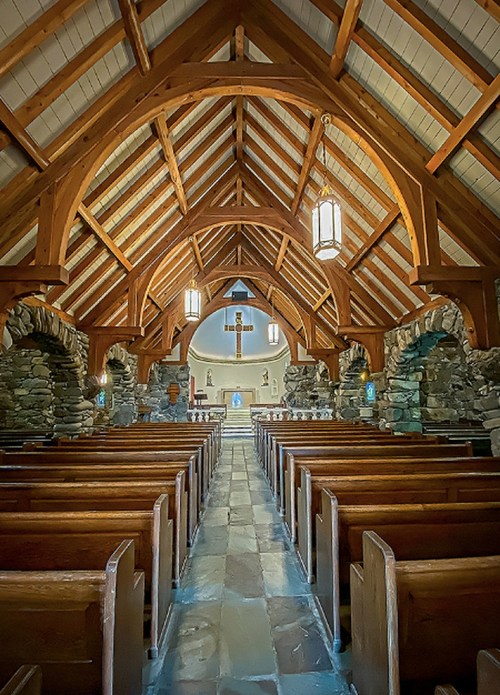 Interior of a rustic church with wooden beams, stone walls, and rows of wooden pews leading to an altar.