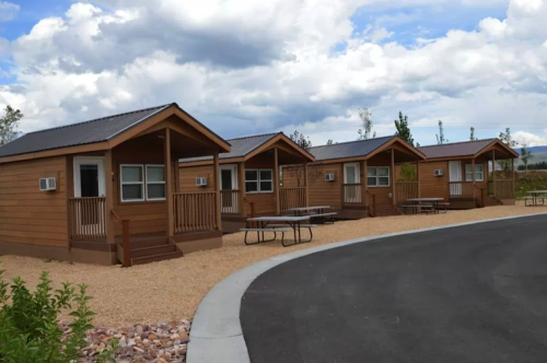 Four wooden cabins with porches and air conditioning units, set along a curved pathway under a cloudy sky.