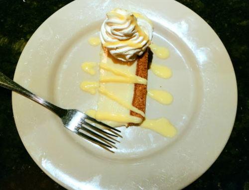 A slice of cake topped with whipped cream and drizzled with a yellow sauce, served on a white plate with a fork.