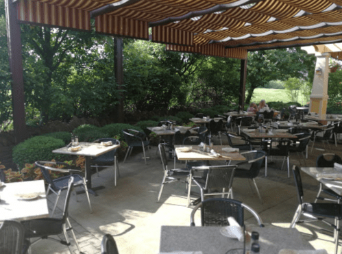 Outdoor dining area with empty tables and chairs under a striped canopy, surrounded by greenery.