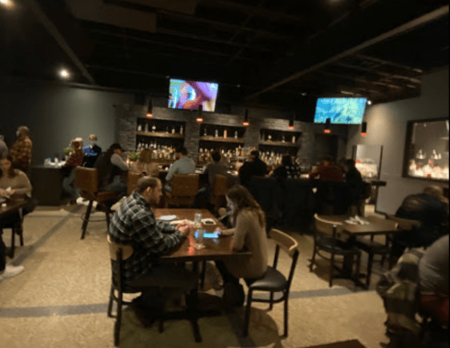 A cozy bar scene with patrons seated at tables and a well-stocked back bar, featuring two TVs showing content.