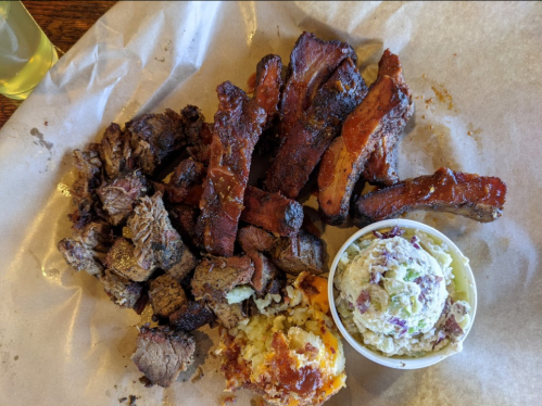 A platter of assorted meats including brisket and ribs, accompanied by a side of potato salad and cornbread.