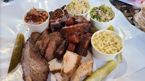 A platter of assorted barbecue meats, including brisket and ribs, served with sides of coleslaw, baked beans, and macaroni.