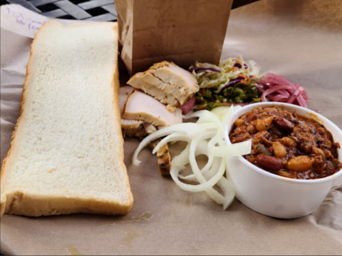 A plate featuring sliced chicken, a bowl of chili, onions, pickled vegetables, and a slice of bread on brown paper.