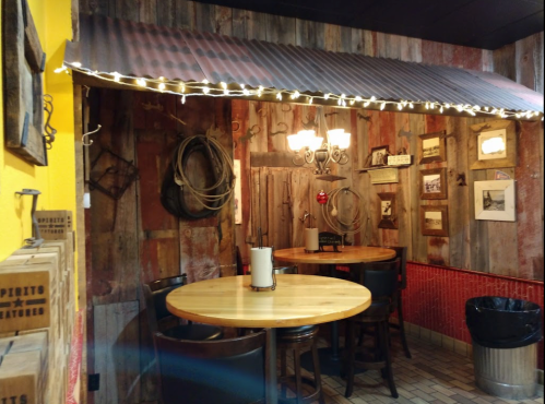Cozy restaurant corner with wooden walls, string lights, and two round tables. Decor includes vintage items and framed photos.
