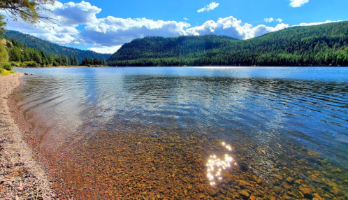 A serene lake surrounded by mountains, with sparkling water and a clear blue sky dotted with clouds.