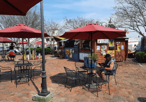 A sunny outdoor café with red umbrellas, tables, and a wooden kiosk surrounded by trees and brick pathways.