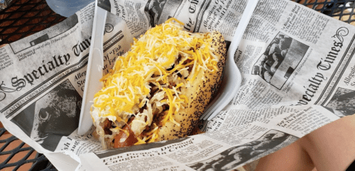 A hot dog topped with shredded cheese and onions, served in a paper tray lined with newspaper.